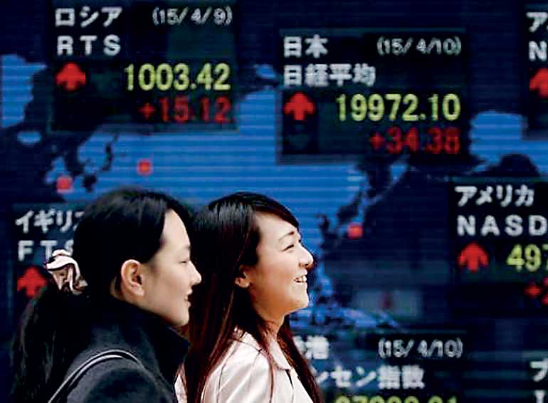 Pedestrians walk past an electronic board showing the stock market indices of various countries outside a brokerage in Tokyo