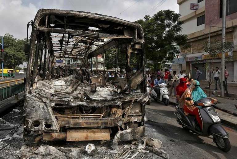 Commuters ride their motocycles next to the wreckage of a bus that was burnt in the clashes between the police and protesters in Ahmedabad