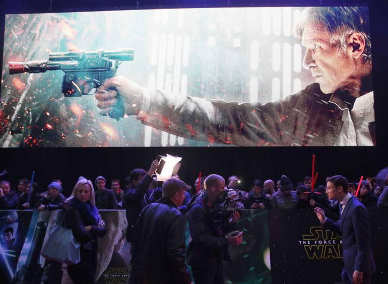 A television presenter does a piece to camera under a giant image of Han Solo, as enthusiasts wait to spot members of the cast at the European Premiere of Star Wars, The Force Awakens in Leicester Square, London